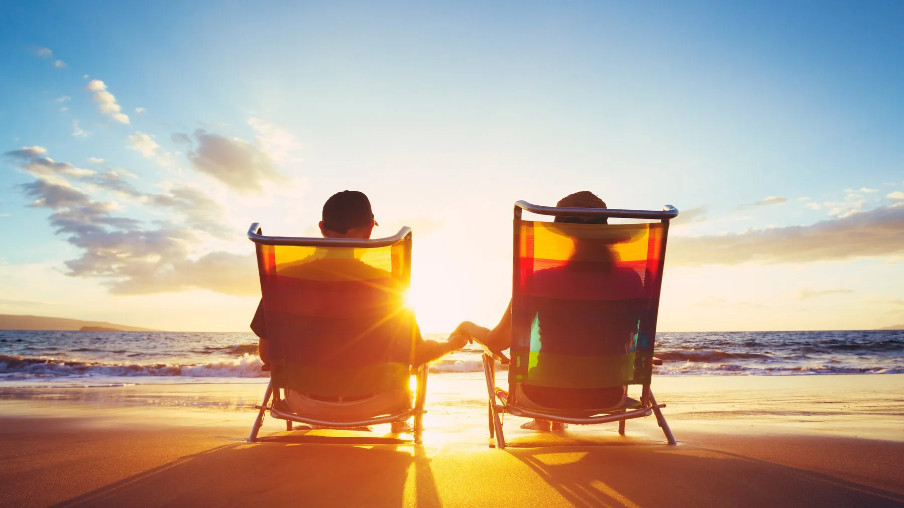 A couple holding hands in beach chairs, watching the sun set over the sea