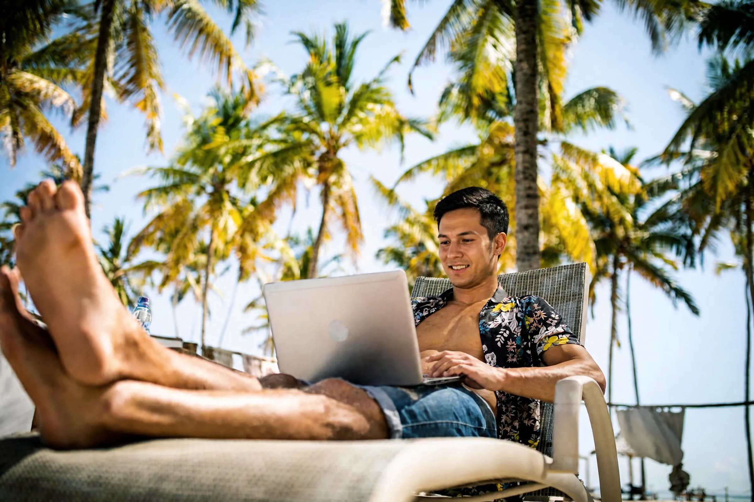 Member working on a laptop from a beach lounger under palm trees